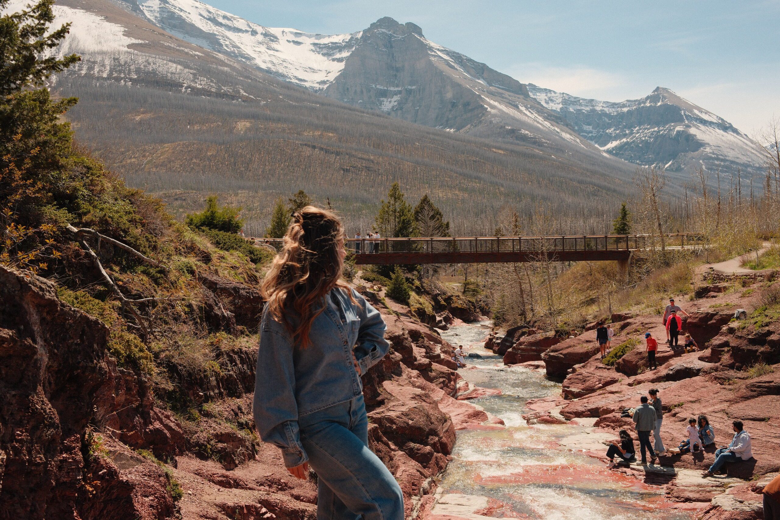 Visitor enjoying the scenic views at Red Rock Canyon in Waterton Lakes National Park, with red rock formations, a mountain stream, and towering Rocky Mountain peaks.