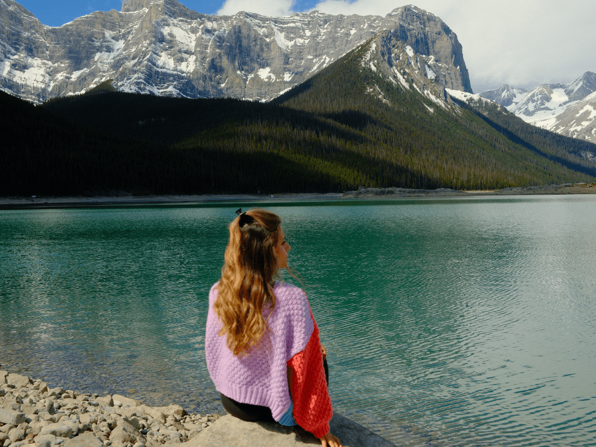 Woman sitting on a rock over looking upper kananskis lake with snow capped, mountain backdrop and blue skis